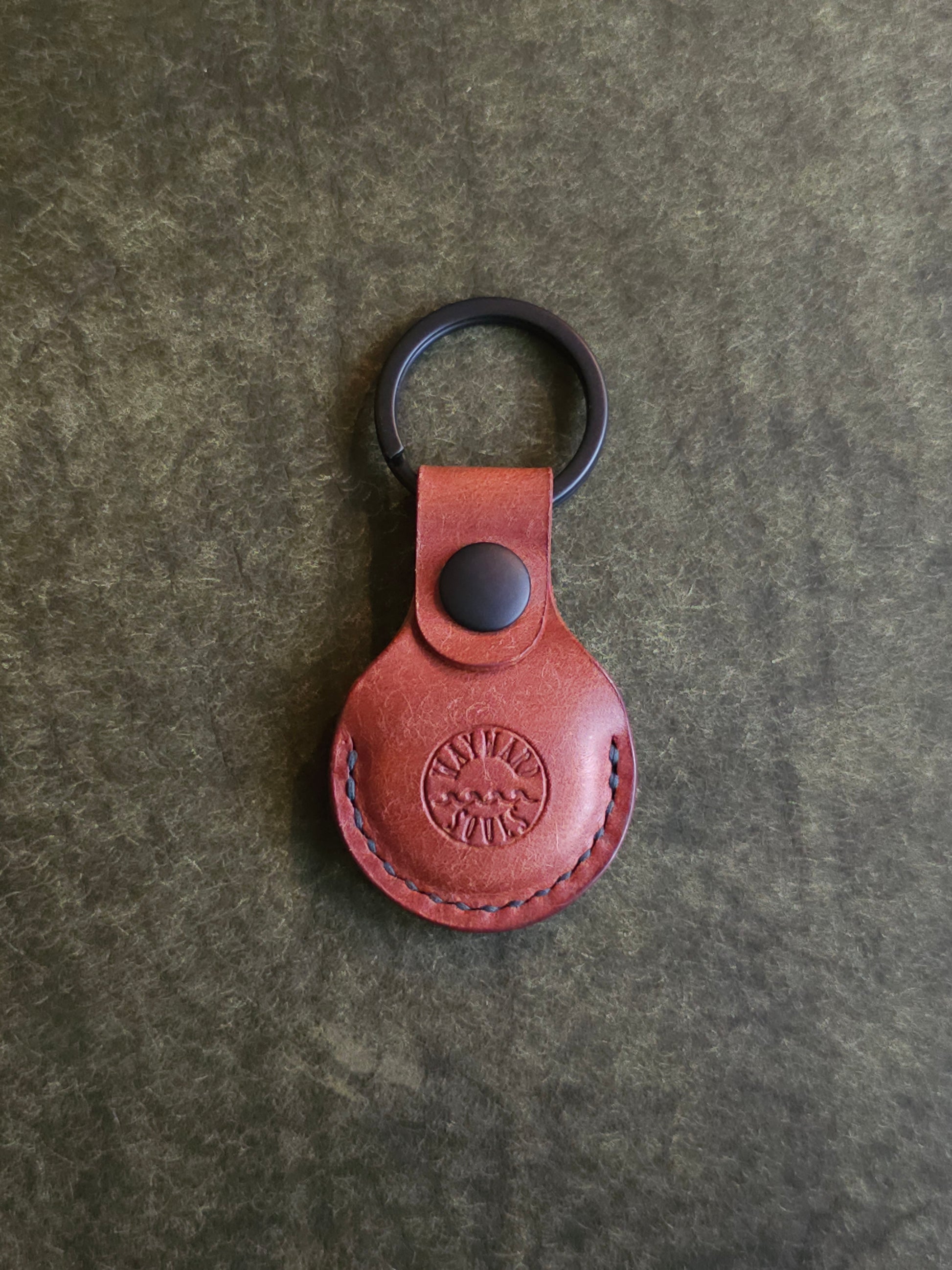 Circular leather air tag holder attached to black key ring. The Wayward Souls logo is stamped into the center.