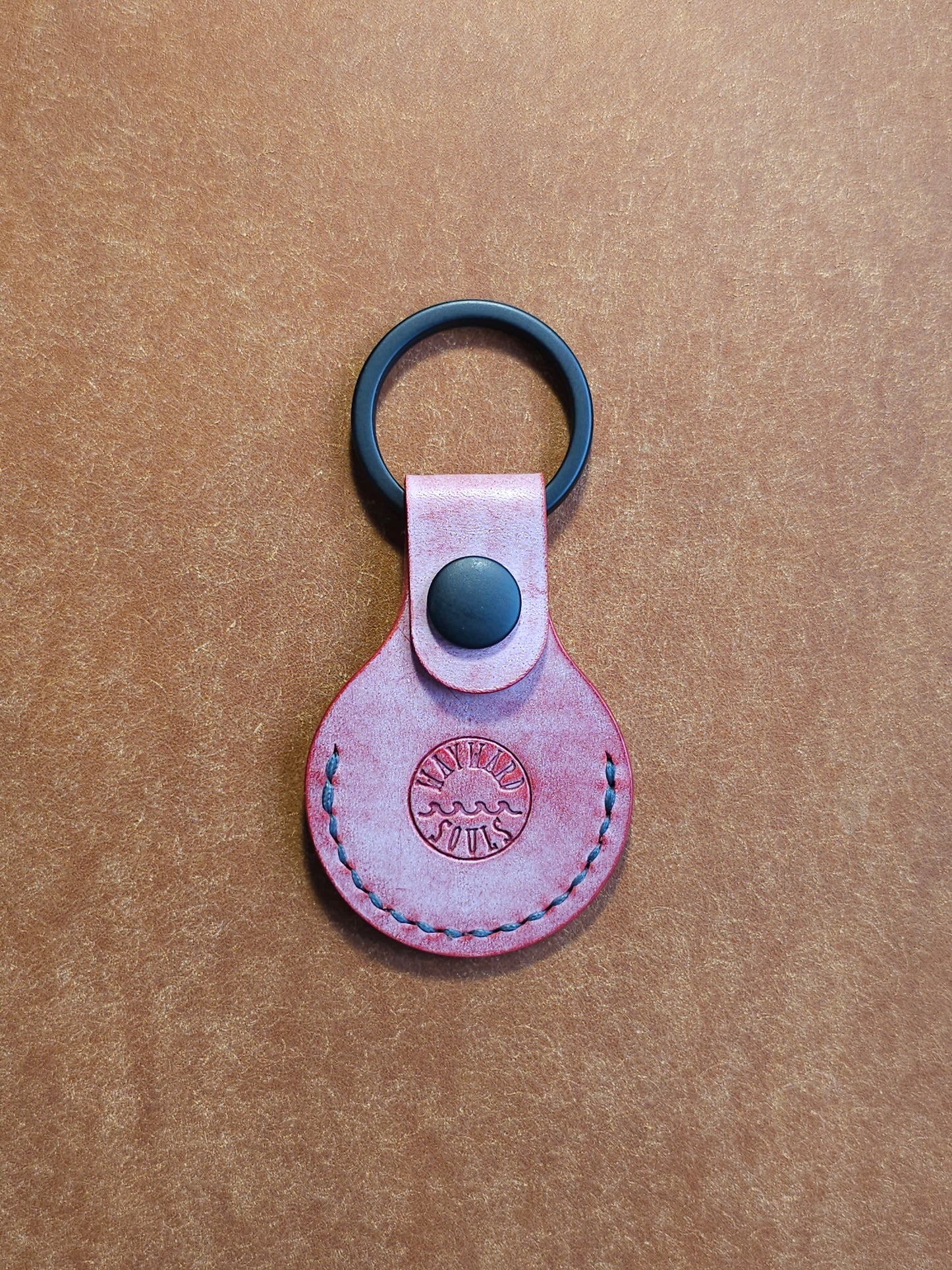  Circular leather air tag holder attached to black key ring. The Wayward Souls logo is stamped into the center.