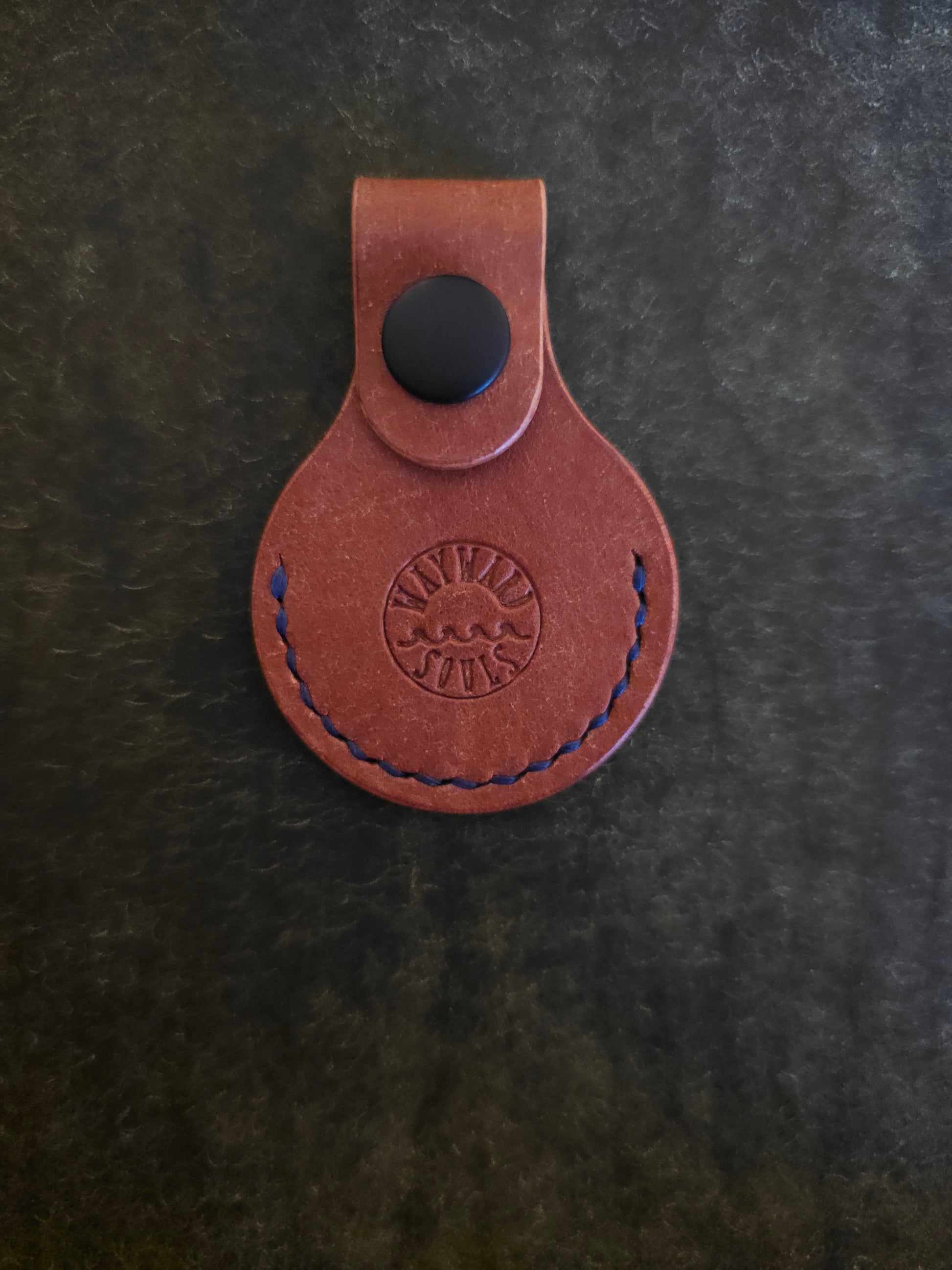 Circular leather air tag holder. The Wayward Souls logo is stamped into the center.